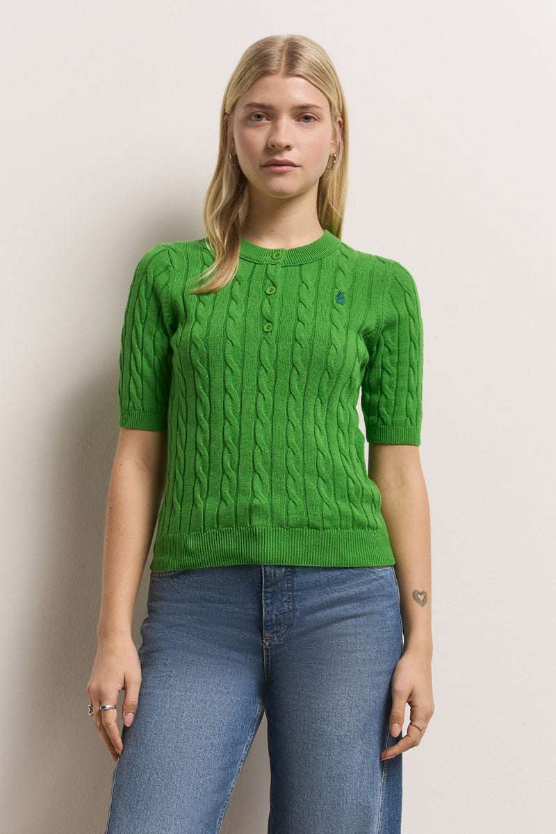 Eine junge Frau mit langen blonden Haaren trägt einen hellgrünen, kurzärmeligen Princess Goes Hollywood Zopfstrick Pullover aus Baumwolle und blaue Jeans. Sie steht vor einem schlichten, gebrochen weißen Hintergrund und hat ein kleines Herz-Tattoo auf ihrem linken Unterarm.