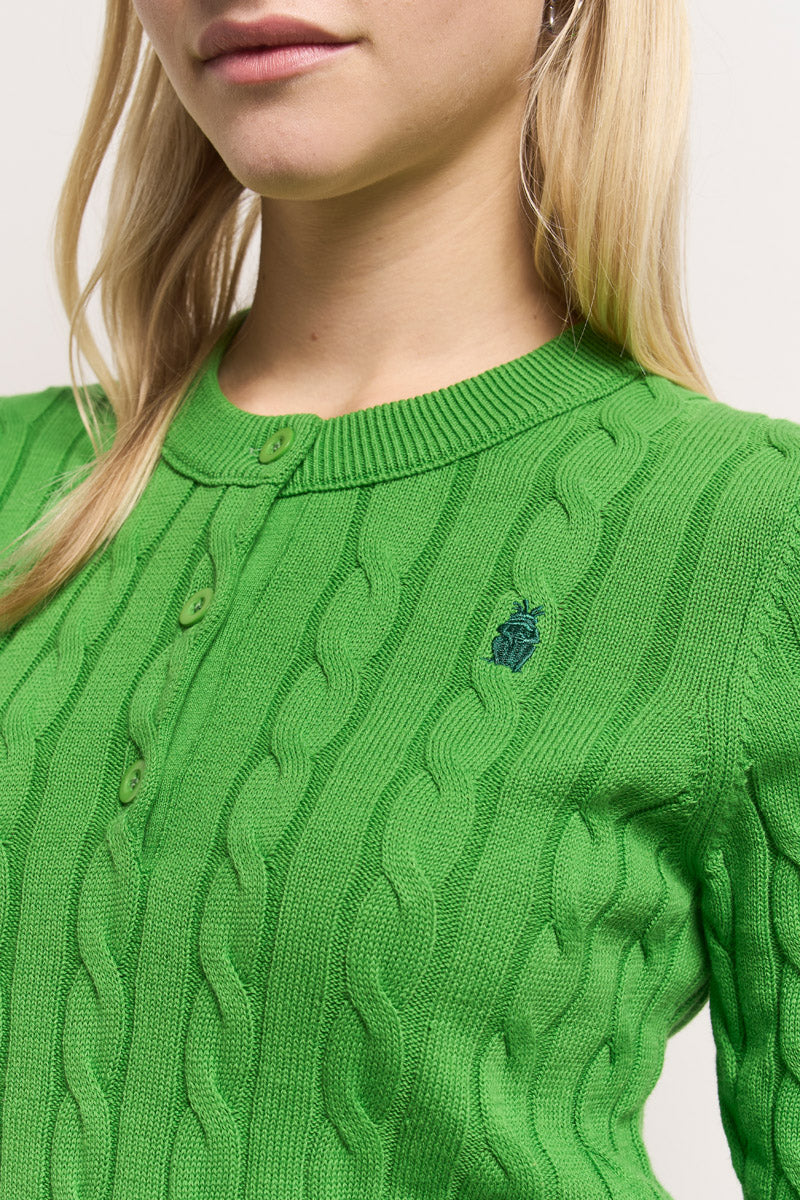 Eine Nahaufnahme zeigt eine Frau in einem leuchtend grünen Princess Goes Hollywood Zopfstrick Pullover aus Baumwolle mit grünen Knöpfen und einer Frosch-Stickerei auf der linken Brust, die ihren Oberkörper, ihr langes blondes Haar und ihre hellrosa Lippen hervorhebt.