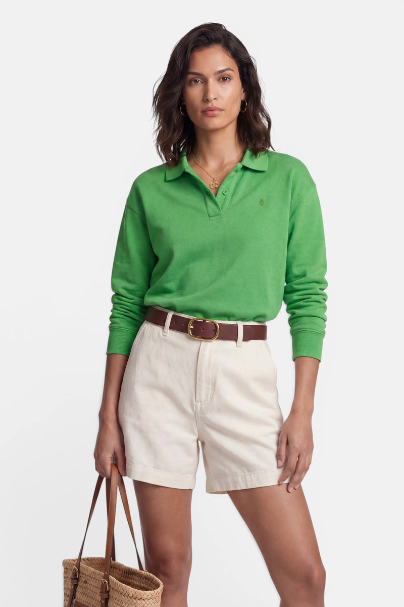 Eine Frau mit welligem braunem Haar trägt ein grünes, langärmeliges Polokragen-Shirt der Marke „Princess Goes Hollywood“, das sie in beigefarbene Shorts mit hoher Taille und einem braunen Gürtel gesteckt hat. Sie hält eine geflochtene Tragetasche in der Hand, trägt eine goldene Halskette als Accessoire und steht vor einem schlichten weißen Hintergrund.