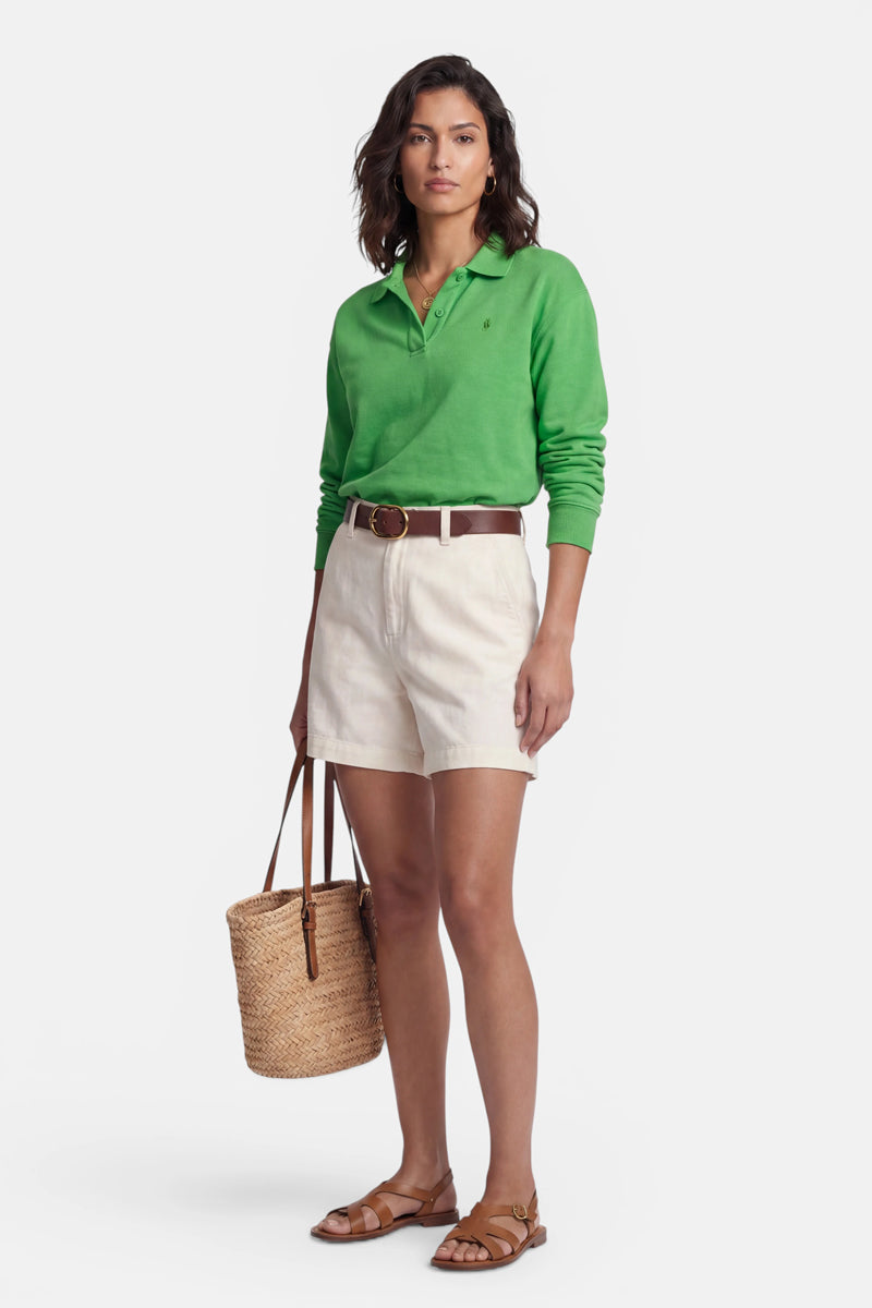 Eine Frau blickt nach vorne. Sie trägt ein hellgrünes Baumwoll-Sweatshirt mit Polokragen der Marke „Princess Goes Hollywood“, beige Shorts mit hoher Taille und einem braunen Gürtel sowie eine geflochtene Strohtasche. Vor einem weißen Hintergrund hat sie offenes dunkles Haar und einen neutralen Gesichtsausdruck.