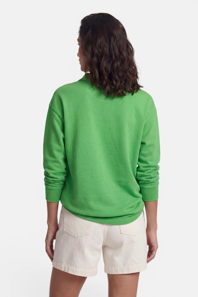 Vor einem schlichten weißen Hintergrund ist eine Person von hinten zu sehen, die welliges, schulterlanges braunes Haar hat und ein hellgrünes „Princess Goes Hollywood“-Baumwoll-Sweatshirt mit Polokragen sowie cremefarbene Shorts mit großen Gesäßtaschen trägt.