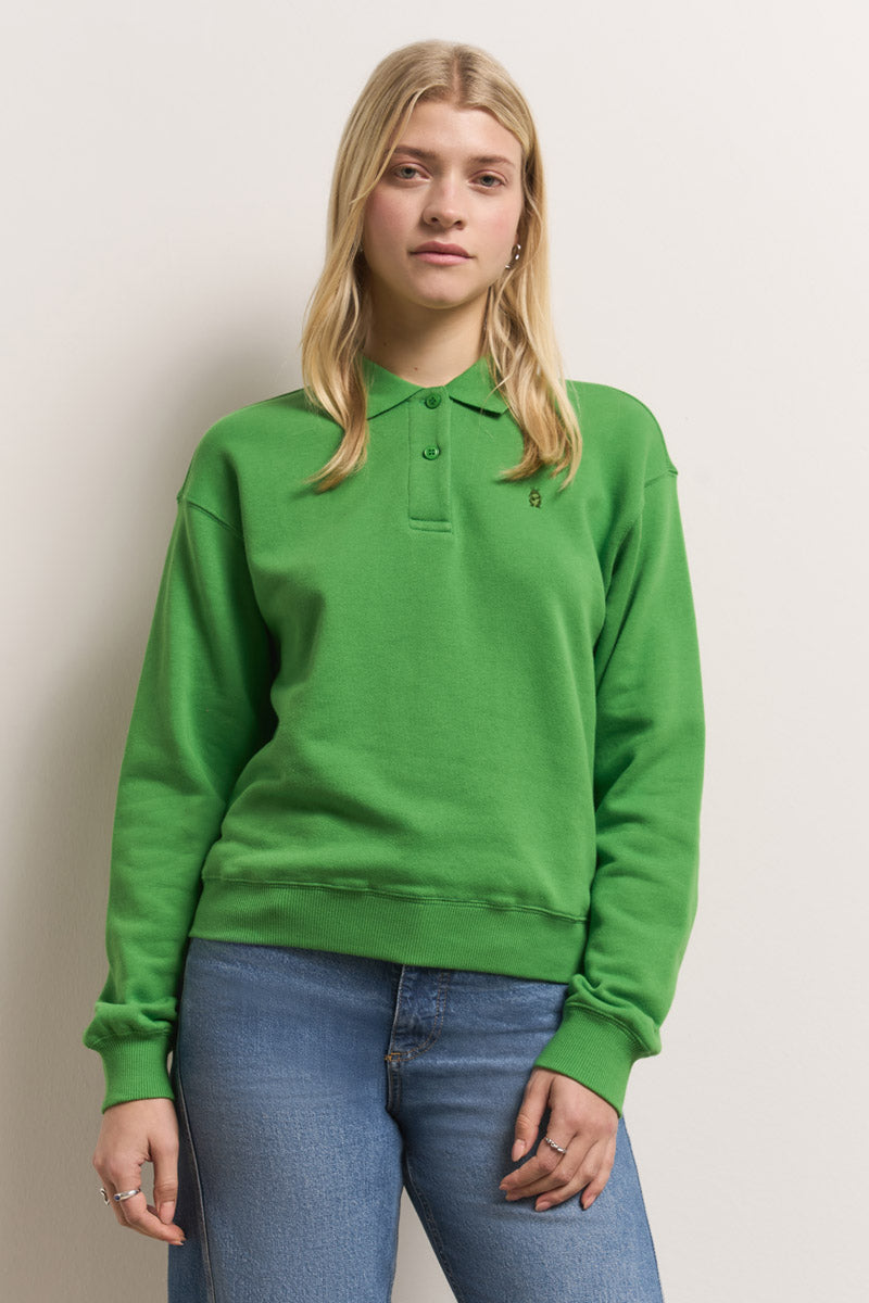 Eine junge Frau mit langen blonden Haaren steht vor einer schlichten Wand. Sie trägt ein hellgrünes Baumwolle Polokragen Sweatshirt von Princess Goes Hollywood und hellblaue Jeans. Ihr Gesichtsausdruck ist neutral, ihre linke Hand ruht an ihrer Seite.