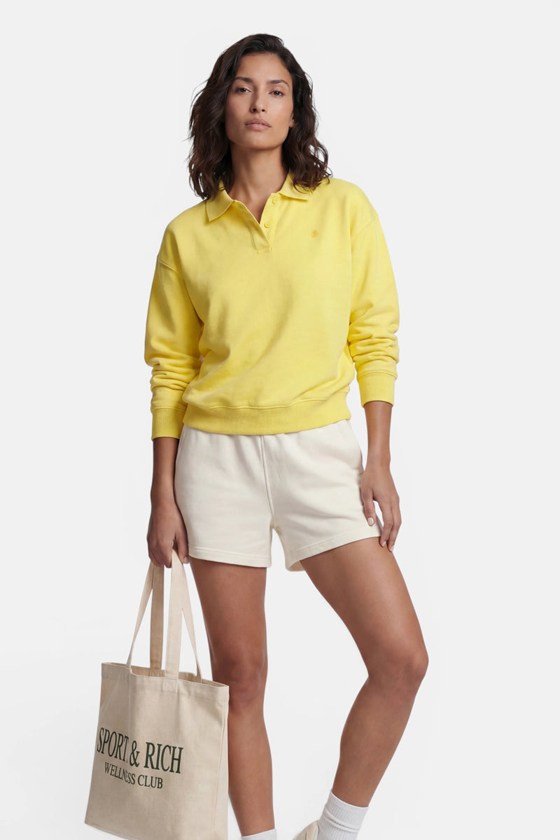 Eine Frau mit schulterlangem braunem Haar steht vor einem weißen Hintergrund. Sie trägt ein gelbes „Princess Goes Hollywood“-Sweatshirt mit Polokragen aus Baumwolle, cremefarbene Shorts und weiße Socken und hält eine beigefarbene Tragetasche des „SPORTY & RICH WELLNESS CLUB“ in der Hand.