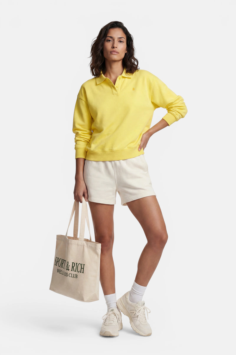 Eine Person steht selbstbewusst vor einem weißen Hintergrund und trägt ein leuchtend gelbes „Princess Goes Hollywood“-Sweatshirt aus Baumwolle mit Polokragen, cremefarbene Shorts, weiße Socken und beige Turnschuhe; in der Hand hält sie eine beige Tragetasche mit der Aufschrift „SPORTY &amp; RICH WELLNESS CLUB“ in Schwarz.