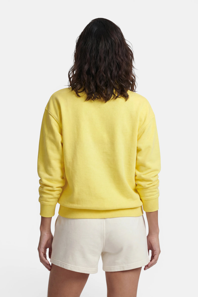 Eine Person mit welligem, schulterlangem braunem Haar ist von hinten zu sehen. Sie trägt ein leuchtend gelbes „Princess Goes Hollywood“-Sweatshirt aus Baumwolle mit Polokragen und cremefarbene Shorts; die Arme liegen entspannt an den Seiten, vor einem schlichten weißen Hintergrund.