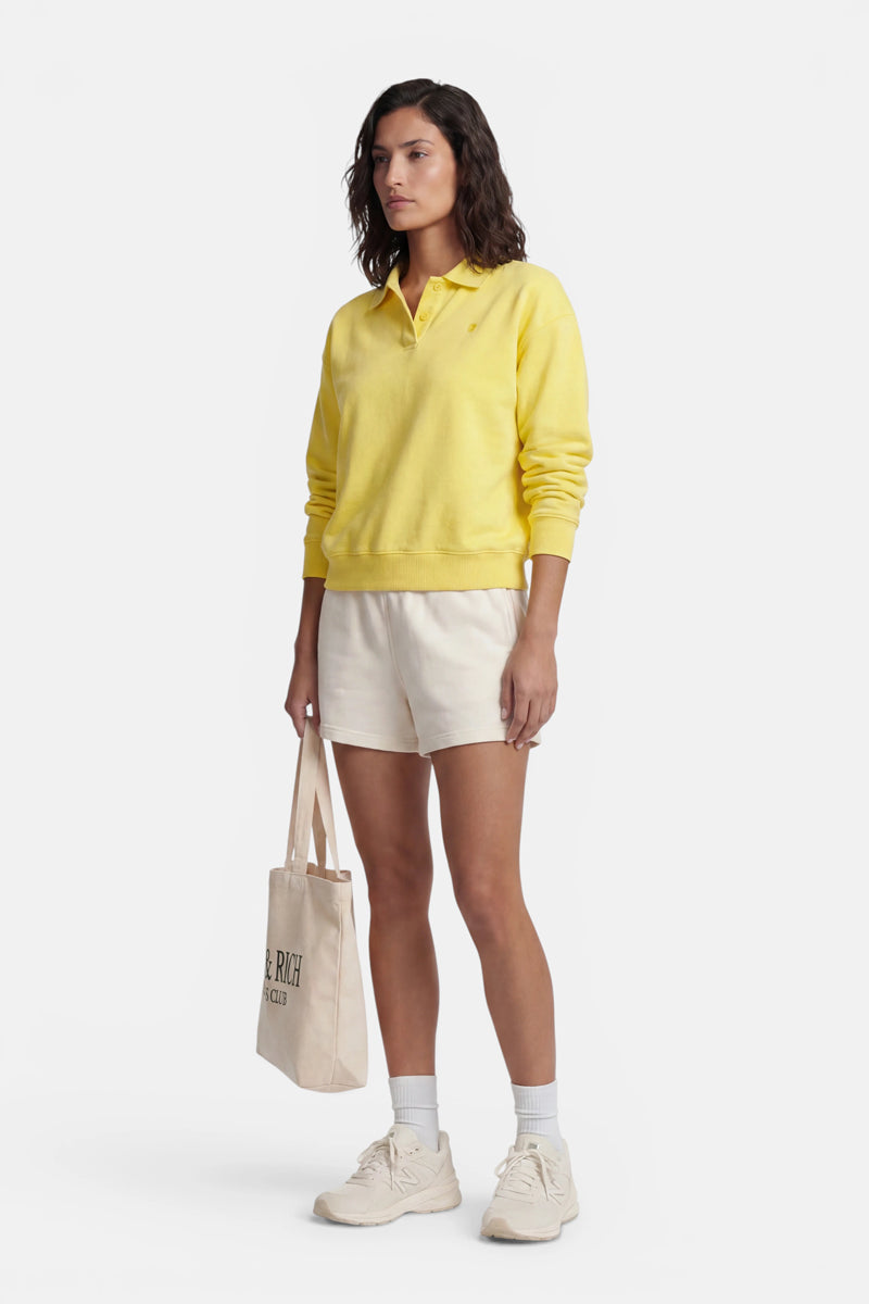 Eine Frau blickt nach vorne. Sie trägt ein gelbes, langärmeliges „Princess Goes Hollywood“-Sweatshirt aus Baumwolle, hellbeige Shorts, weiße Socken und beige Turnschuhe und hält eine beige Tragetasche in der Hand. Der Hintergrund ist schlicht weiß.