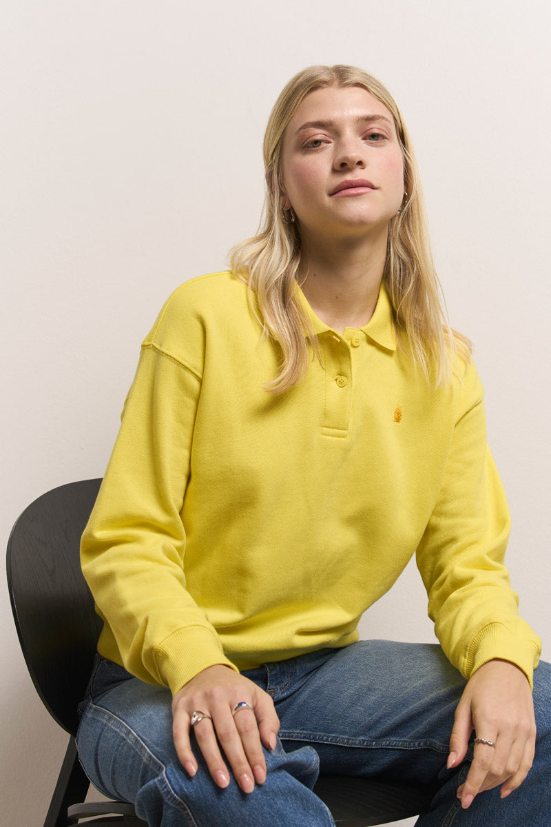 Eine Person mit langen blonden Haaren trägt ein gelbes Princess Goes Hollywood Polokragen-Sweatshirt aus Baumwolle und blaue Jeans. Sie sitzt entspannt auf einem schwarzen Stuhl vor einem schlichten hellen Hintergrund, eine Hand auf dem Knie, die andere auf dem Stuhl.