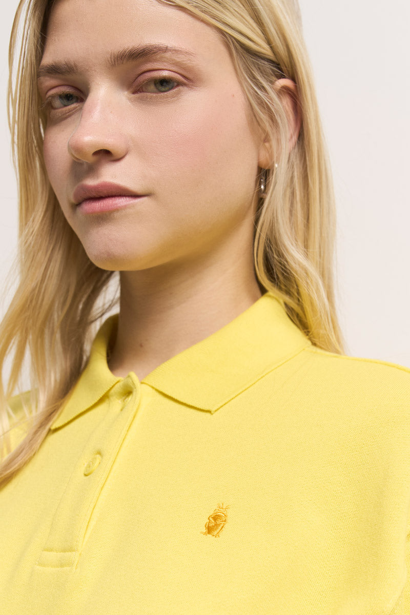 Eine junge Frau mit langen blonden Haaren und heller Haut trägt ein leuchtend gelbes Baumwoll-Poloshirt von Princess Goes Hollywood mit einem kleinen gestickten Logo auf der Brust. Sie ist dezent rosa geschminkt und blickt vor einem hellen Hintergrund leicht an der Kamera vorbei.