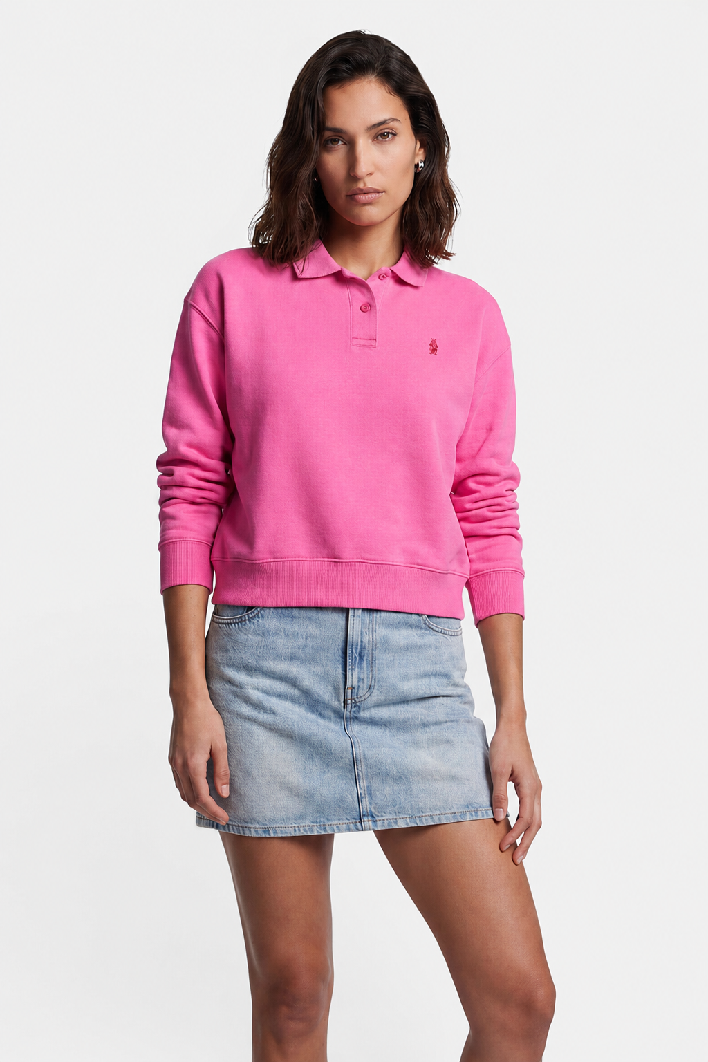 Eine Person mit schulterlangem braunem Haar trägt ein leuchtend pinkes Baumwoll-Sweatshirt mit Polokragen von „Princess Goes Hollywood“, dazu einen hellblauen Jeans-Minirock. Sie steht vor einem schlichten weißen Hintergrund, mit ausdruckslosem Gesicht und einer Hand auf dem Oberschenkel.