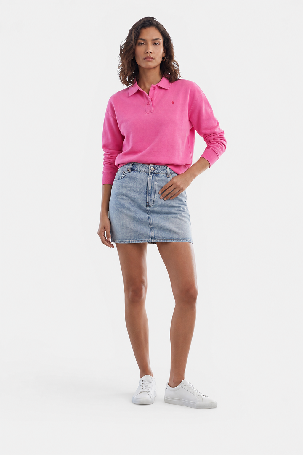 Eine Frau steht mit dem Blick nach vorne und trägt ein leuchtend pinkes Langarm-Sweatshirt mit Polokragen von „Princess Goes Hollywood“, das sie in einen hellblauen Jeans-Minirock gesteckt hat. Sie hat kurzes, welliges Haar, trägt weiße Sneaker und blickt mit ausdruckslosem Gesicht in die Kamera, während sie vor einem schlichten weißen Hintergrund posiert.