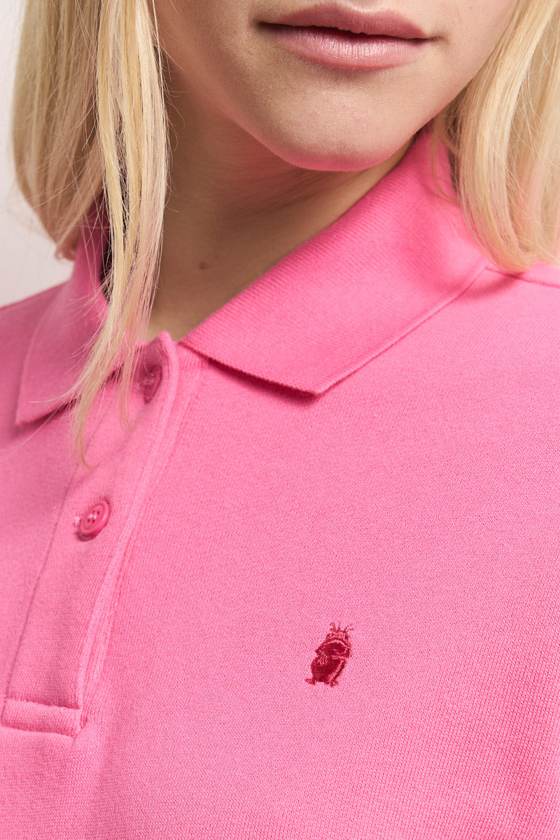 Nahaufnahme einer Person mit hellblondem Haar, die ein hellrosa Princess Goes Hollywood Baumwolle-Poloshirt mit rosa Knöpfen, einem gestickten roten Tierlogo auf der Brust und einem klassischen Polokragen trägt. Nur der untere Teil des Gesichts und der obere Teil des Oberkörpers sind sichtbar.