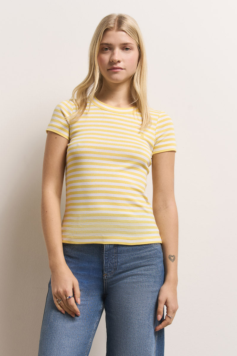 Eine junge Frau mit langen blonden Haaren trägt ein kurzärmeliges, gelb-weiß gestreiftes Baumwoll-T-Shirt von Princess Goes Hollywood und blaue Jeans. Sie steht an einer gebrochen weißen Wand und schaut mit neutralem Blick in die Kamera.