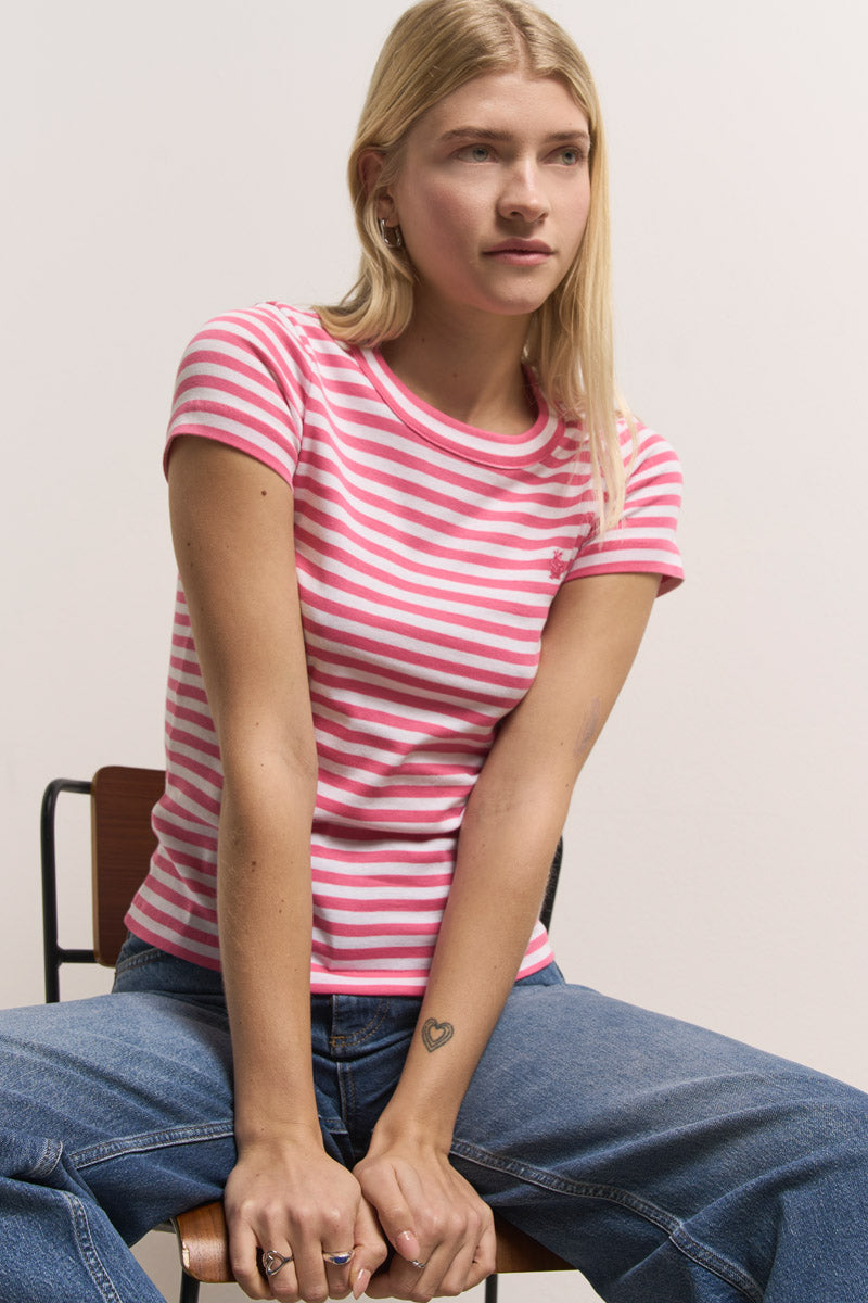 Eine junge Frau mit glattem blondem Haar sitzt auf einem Stuhl und trägt ein rosa-weiß gestreiftes Baumwoll-T-Shirt von Princess Goes Hollywood und blaue Jeans. Ihre Hände ruhen auf den Knien und zeigen eine kleine Herz-Tätowierung auf ihrem rechten Handgelenk. Der Hintergrund ist hell.