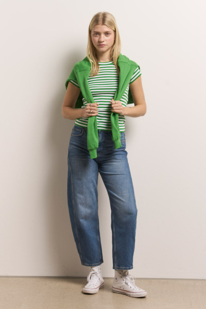 Eine junge Frau mit glattem blondem Haar steht vor einer schlichten Wand. Sie trägt ein Princess Goes Hollywood gestreiftes Baumwoll-T-Shirt, ein grünes Sweatshirt mit Frosch-Stickerei über den Schultern, weite Bluejeans und weiße High-Top-Sneakers und wirkt entspannt.