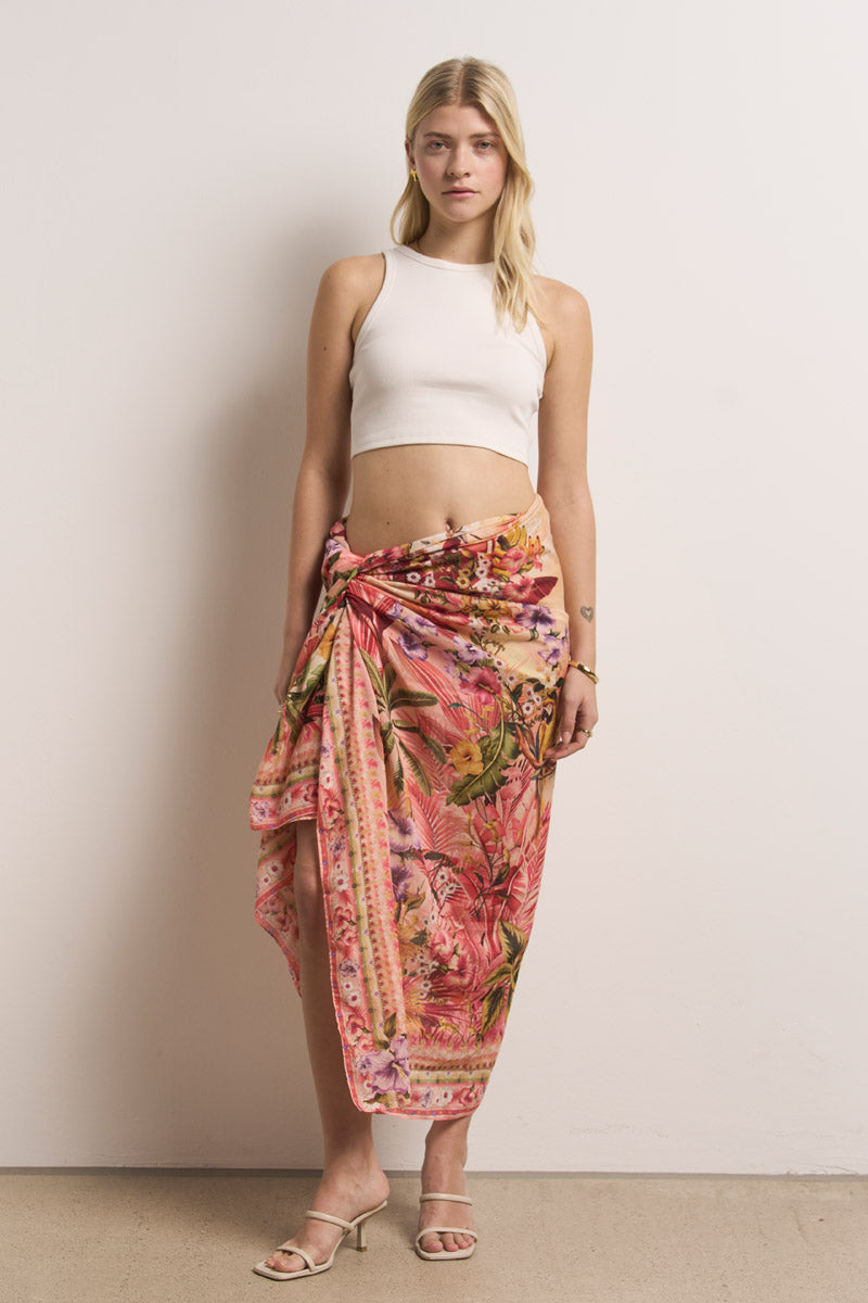 Eine Frau mit langen blonden Haaren steht an einer schlichten Wand. Sie trägt ein tailliertes weißes Crop-Top und einen rosafarbenen Pareo mit tropischem Muster von Princess Goes Hollywood, dazu Sandalen mit offenen Absätzen. Ihr Gesichtsausdruck ist neutral und ihre Arme ruhen entspannt an ihren Seiten.