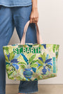 Eine Person in blauen Jeans und blauem Hemd hält eine farbenfrohe Princess Goes Hollywood Strandtasche aus Baumwolle mit beigen Griffen, auf denen grüne tropische Blätter, blaue Vasen und die fetten Worte ST. BARTH und CARIBBEAN.