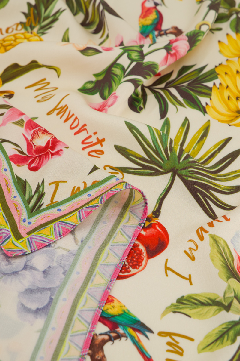 Eine Nahaufnahme des Princess Goes Hollywood-Stofftuchs zeigt lebhafte tropische Prints - Papageien, Palmenblätter, Bananen und Orchideen. Goldfarbene, kursive Akzente und eine mehrfarbige, geometrische Kante machen dieses Accessoire zu einem echten Styling-Highlight.