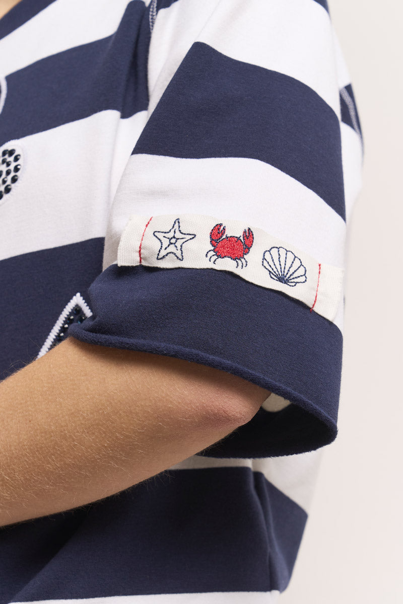 Eine Nahaufnahme eines marineblau-weiß gestreiften kurzärmeligen Hemds von Princess Goes Hollywood zeigt Ärmelaufschläge mit aufgestickten blauen Seesternen, roten Krabben und weißen Muscheln. Der Arm der Trägerin erscheint vor einem schlichten hellen Hintergrund.