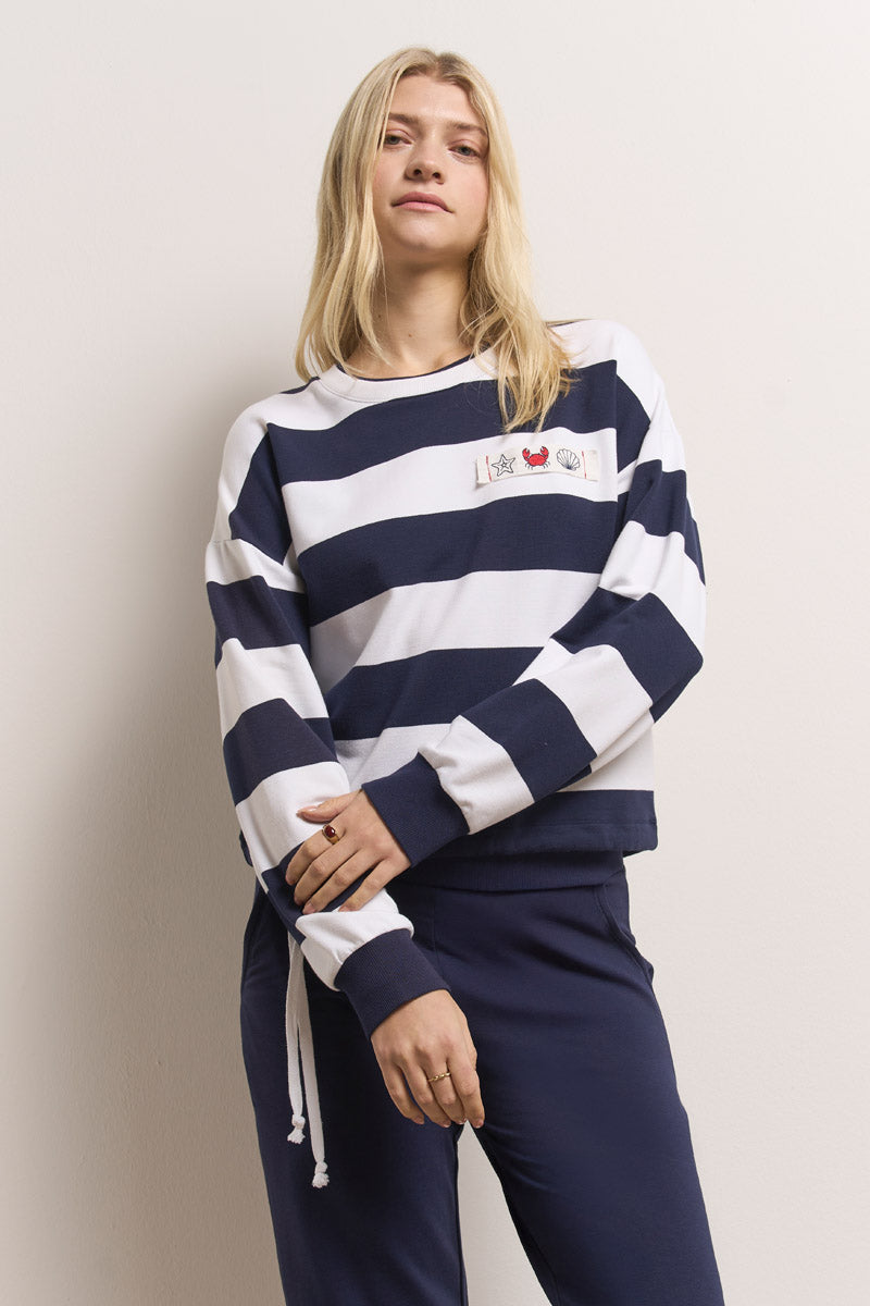 Eine junge Frau mit langen blonden Haaren modelliert ein Princess Goes Hollywood gestreiftes Sweatshirt mit Rundhalsausschnitt und kleiner Bruststickerei. Sie posiert selbstbewusst vor einer cremefarbenen Wand, eine Hand auf der Hüfte, und schaut in die Kamera.