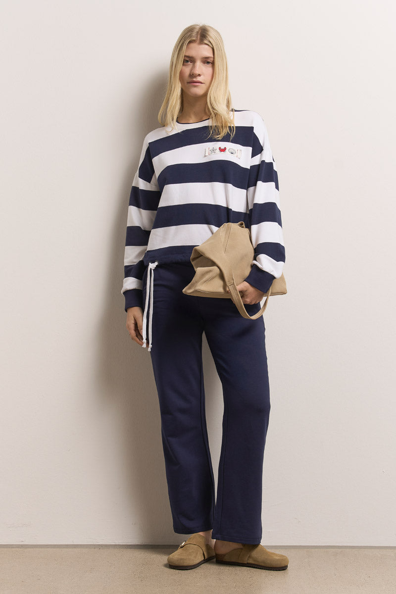 Eine Frau mit glattem blondem Haar steht vor einer schlichten Wand. Sie trägt ein marineblau-weiß gestreiftes Sweatshirt mit Rundhalsausschnitt von Princess Goes Hollywood, eine marineblaue Hose mit Kordelzug und elastischem Saum, hellbraune Slipper und eine hellbraune Tasche, die einen lässigen Look vermittelt.