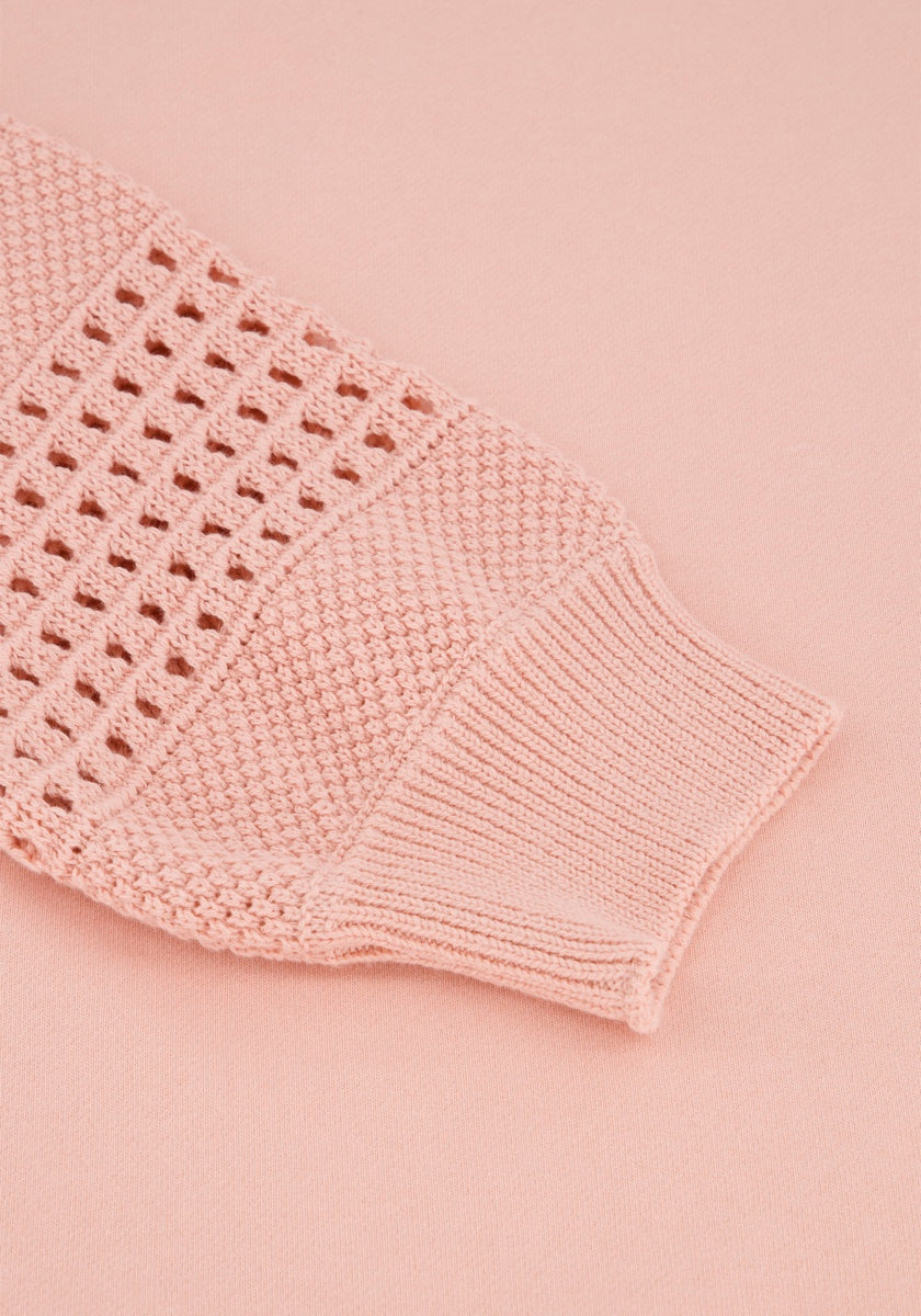 Eine Nahaufnahme eines hellrosa Princess Goes Hollywood-Strickpulloverärmels mit Rippen-, Waben- und offenen Lochstickerei-ähnlichen Mustern, flach auf einem passenden rosa Hintergrund gelegt.
