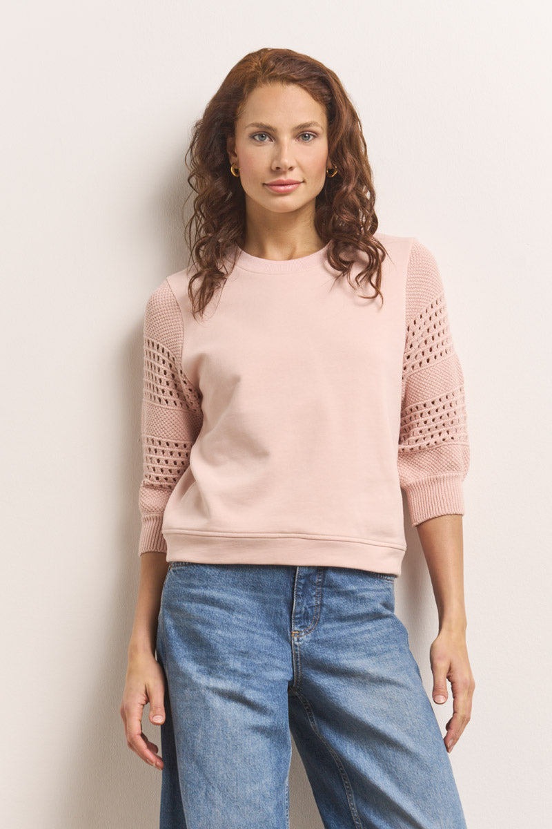 Eine Frau mit gewelltem, rötlich-braunem Haar und goldenen Ohrringen steht vor einer schlichten, hellen Wand. Sie trägt ein hellrosa Baumwolle-Sweatshirt mit Lochstickerei-Einsätzen und eine locker sitzende Bluejeans und blickt selbstbewusst in die Kamera.