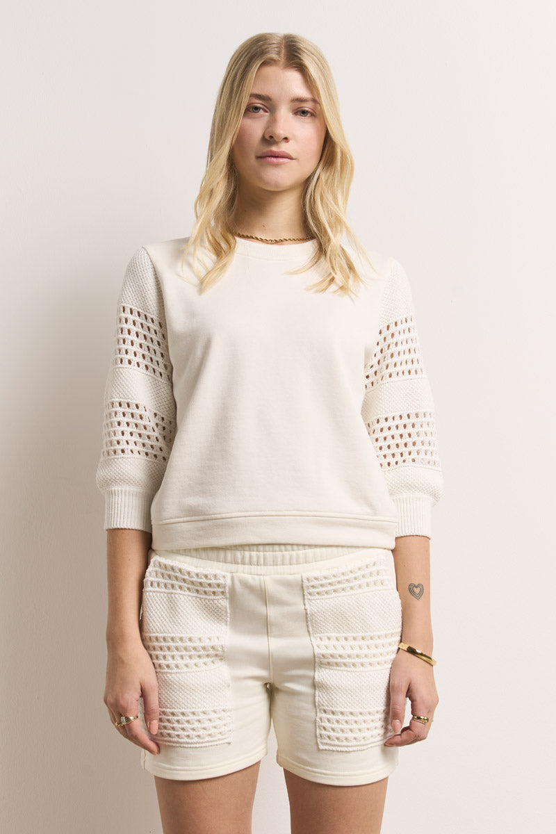 Eine junge Frau mit langen blonden Haaren steht vor einer schlichten weißen Wand. Sie trägt ein cremefarbenes Princess Goes Hollywood-Baumwolloberteil mit gehäkelten Strickärmeln und dazu passende hochtaillierte Shorts mit strukturierten Strickeinsätzen, die mit Ringen, einem Armband und einem kleinen Tattoo verziert sind.