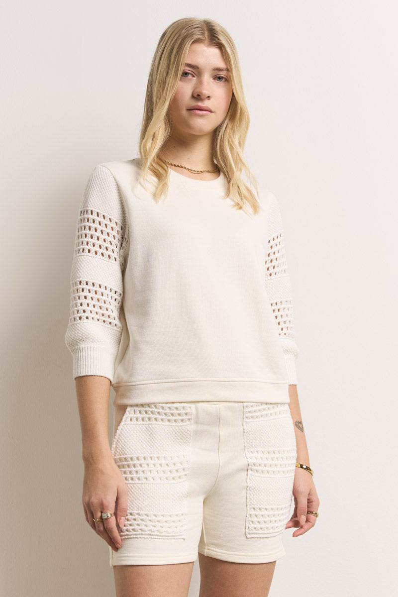 Eine junge Frau mit langen blonden Haaren trägt ein weißes Baumwoll-Strickoberteil mit Lochstickerei-Details an den Ärmeln und dazu passende hochgeschnittene Shorts und steht selbstbewusst vor einem schlichten hellen Hintergrund.