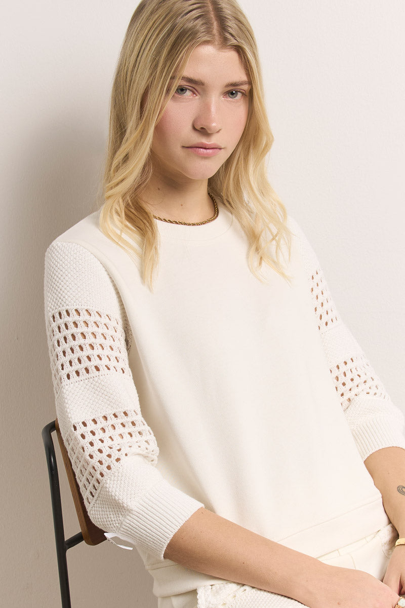 Eine junge Frau mit langen blonden Haaren sitzt auf einem Stuhl vor einer hellen Wand und blickt ruhig in die Kamera. Sie trägt ein Princess Goes Hollywood Sweatshirt Damen aus Baumwolle mit Lochstickerei-Details an den Ärmeln, die Hände ruhen in ihrem Schoß.