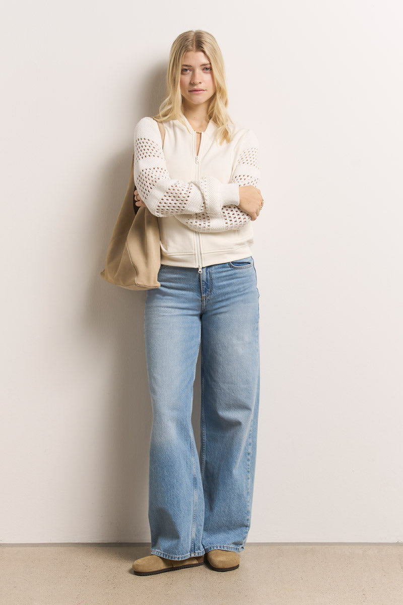 Eine ruhige Frau mit langen blonden Haaren steht mit verschränkten Armen vor einer hellen Wand. Sie trägt eine Princess Goes Hollywood weiße Baumwolle Sweatjacke mit Lochstickerei, hellblaue Jeans mit weitem Bein, hellbraune Wildlederschuhe und eine beigefarbene Umhängetasche.