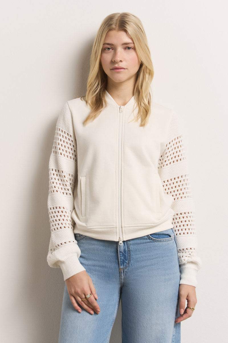 Eine Frau mit langen blonden Haaren trägt eine cremefarbene Princess Goes Hollywood Baumwolle Sweatjacke mit gestrickten Lochstickereiärmeln und hellblaue Jeans. Sie steht vor einer cremefarbenen Wand und blickt neutral nach vorne, die Hände an den Seiten.