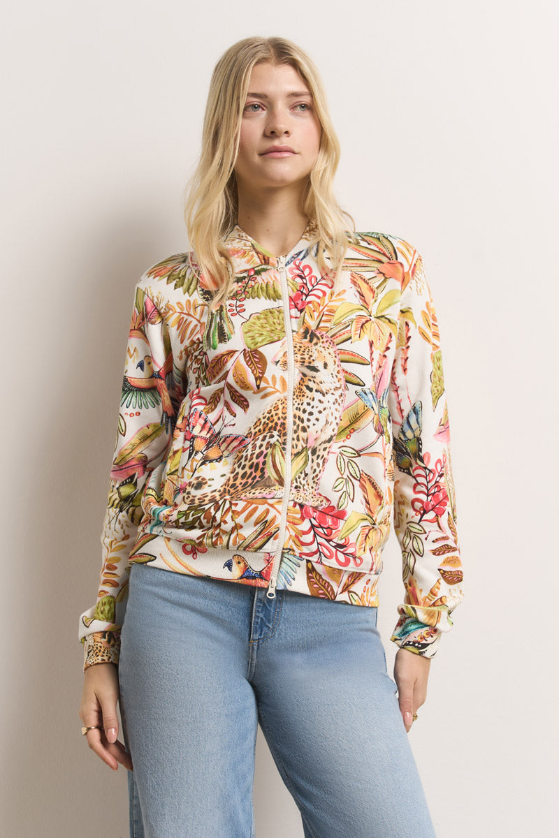 Eine Frau mit langen blonden Haaren trägt eine Princess Goes Hollywood Sweatjacke aus Baumwolle mit Dschungeldruck und Vögeln, Blättern und Blumen. Sie trägt dazu eine hellblaue Jeans und steht vor einem unifarbenen Hintergrund und schaut entspannt zur Seite.