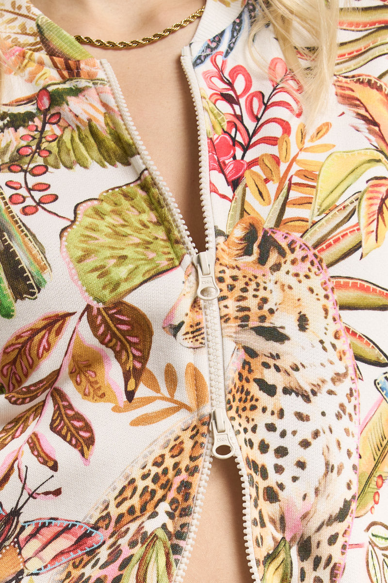 Eine Nahaufnahme einer teilweise geöffneten Princess Goes Hollywood Sweatjacke mit einem auffälligen Dschungelprint aus Blättern, Pflanzen und einem Geparden. Eine goldene Halskette liegt auf der nackten Haut, die durch den geöffneten Reißverschluss sichtbar ist.
