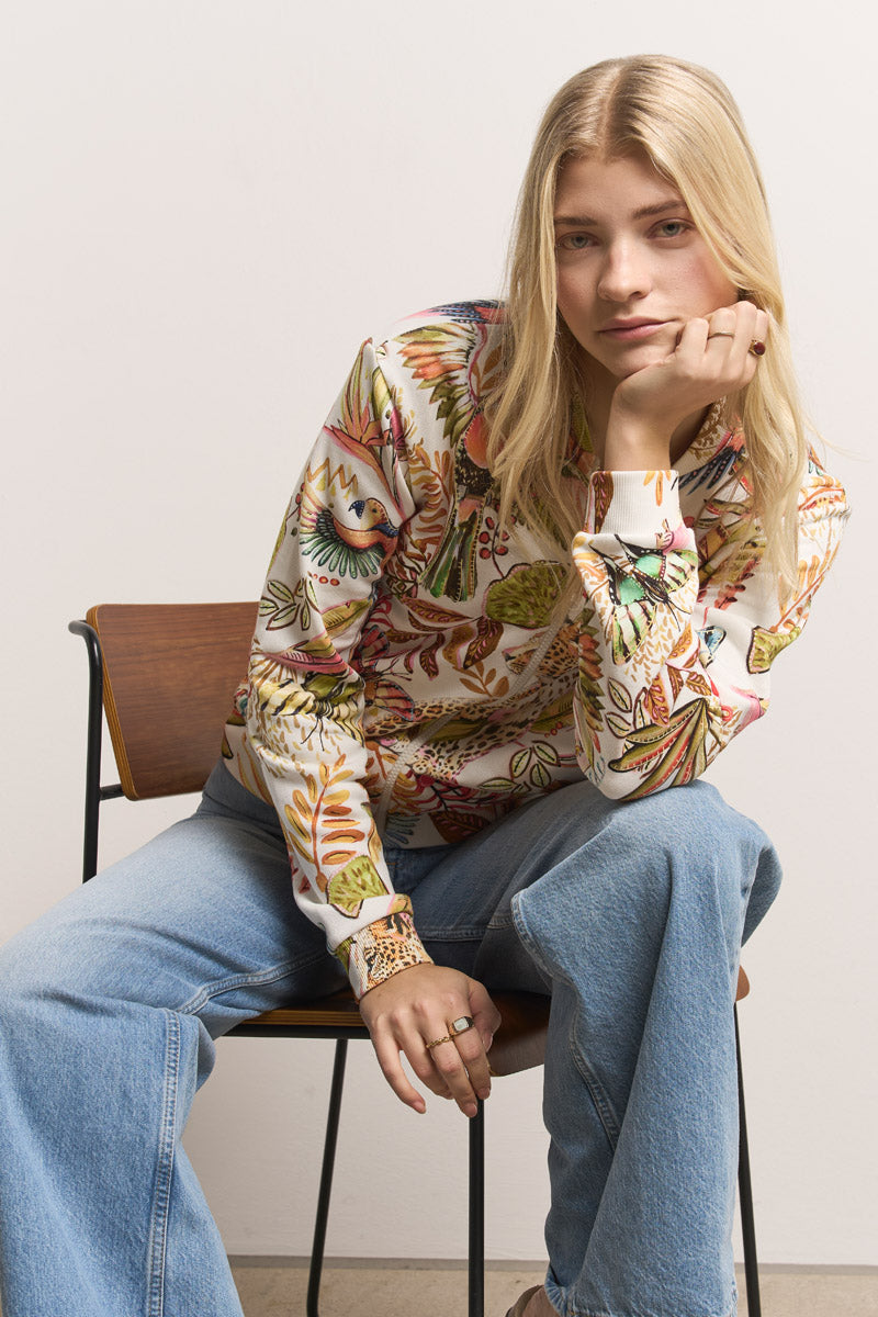 Eine Frau mit glattem blondem Haar sitzt auf einem Stuhl und stützt ihr Kinn auf ihre Hand. Sie trägt eine bunte Baumwoll-Sweatjacke mit Dschungelmuster und blaue Schlagjeans von Princess Goes Hollywood vor einem schlichten, hellen Hintergrund.