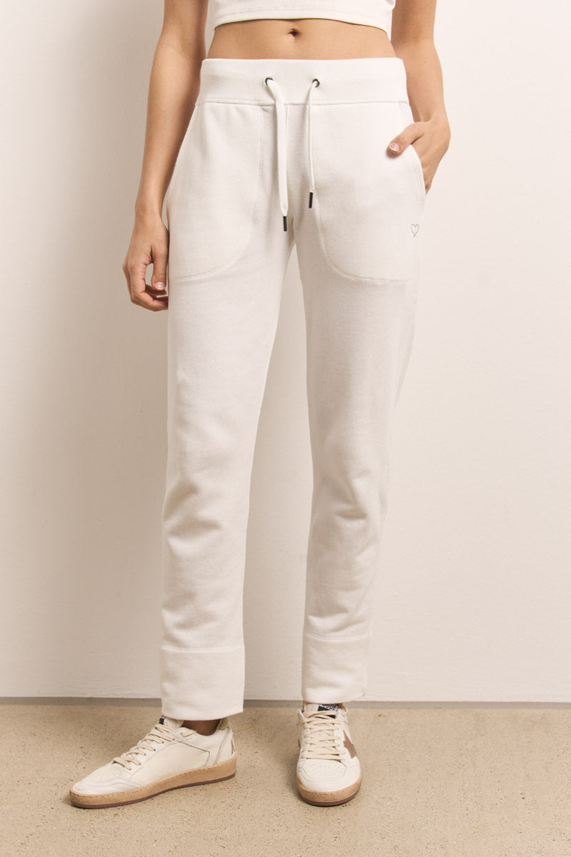 Eine Person, die von der Taille abwärts sichtbar ist, steht an einer hellen Wand und trägt Princess Goes Hollywood, eine cremefarbene Baumwoll-Sweathose mit Vordertaschen und ein passendes Crop-Top. Ihre rechte Hand steckt in einer Tasche, und sie trägt cremefarbene Turnschuhe mit beigen Sohlen.