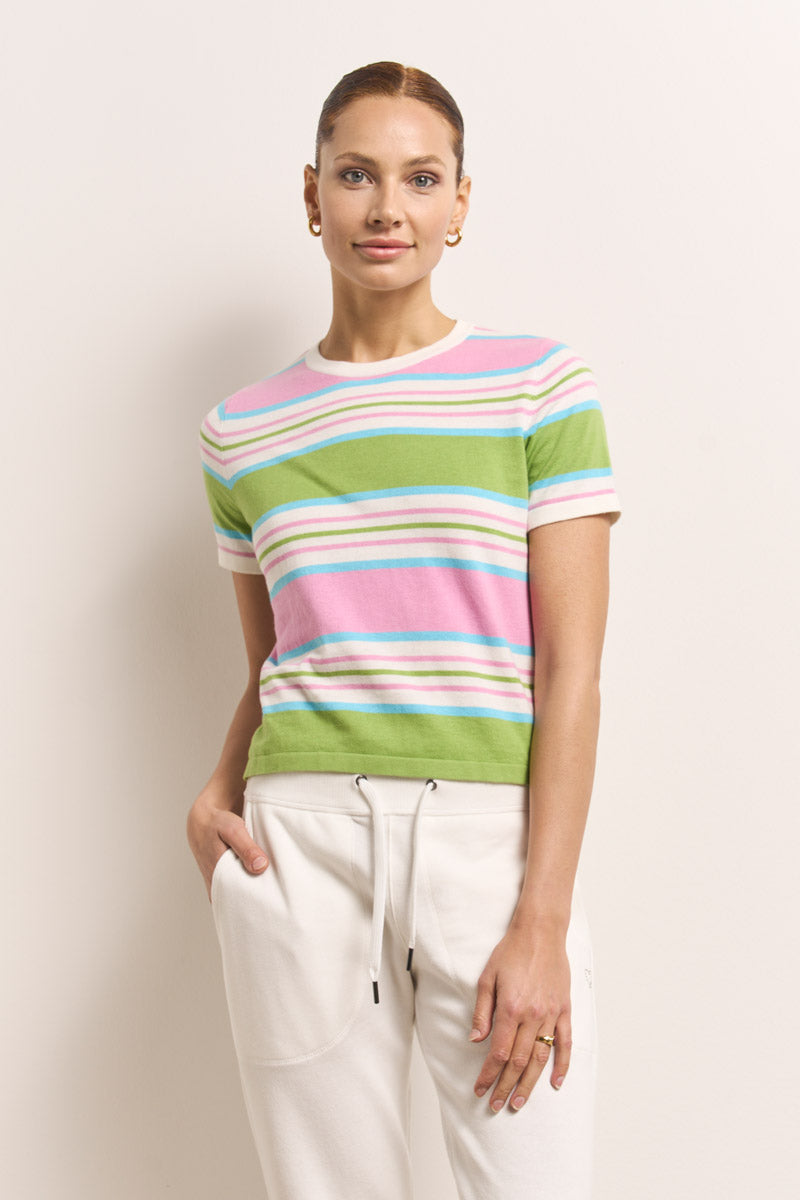 Eine Frau posiert vor einem hellen Hintergrund in einem kurzärmeligen, gestreiften Strickoberteil in Grün, Rosa und Weiß von Princess Goes Hollywood, gepaart mit einer weißen Baumwoll-Sweatpants und goldenen Ohrringen, die linke Hand lässig in der Tasche.