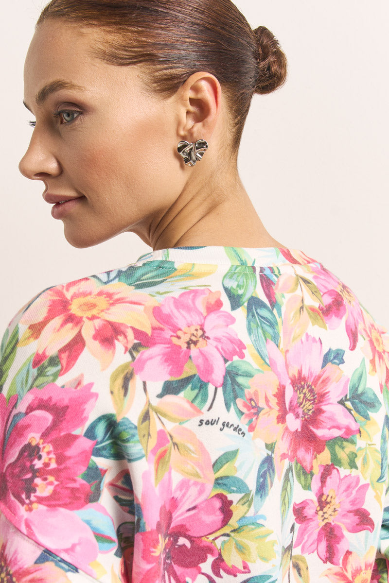Eine Frau mit heller Haut und braunem Haar im Dutt trägt silberne Blumenohrringe und einen weißen Princess Goes Hollywood-Sweatpullover aus Baumwolle mit einem farbenfrohen Blumenaufdruck - rosa und gelbe Blumen, grüne Blätter und "Soul Garden" auf dem Rücken.