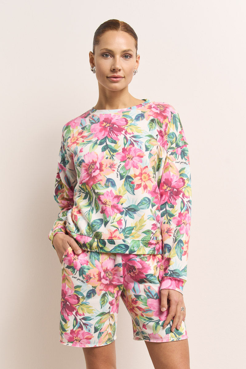 Eine Frau steht an einer hellbeigen Wand und trägt ein Princess Goes Hollywood Blumenprint-Set mit leuchtend rosa, gelben und grünen Blumen auf weißer Baumwolle. Sie blickt nach vorne, eine Hand in der Tasche, ihr langärmeliges Oberteil ähnelt einem Sweatshirt.