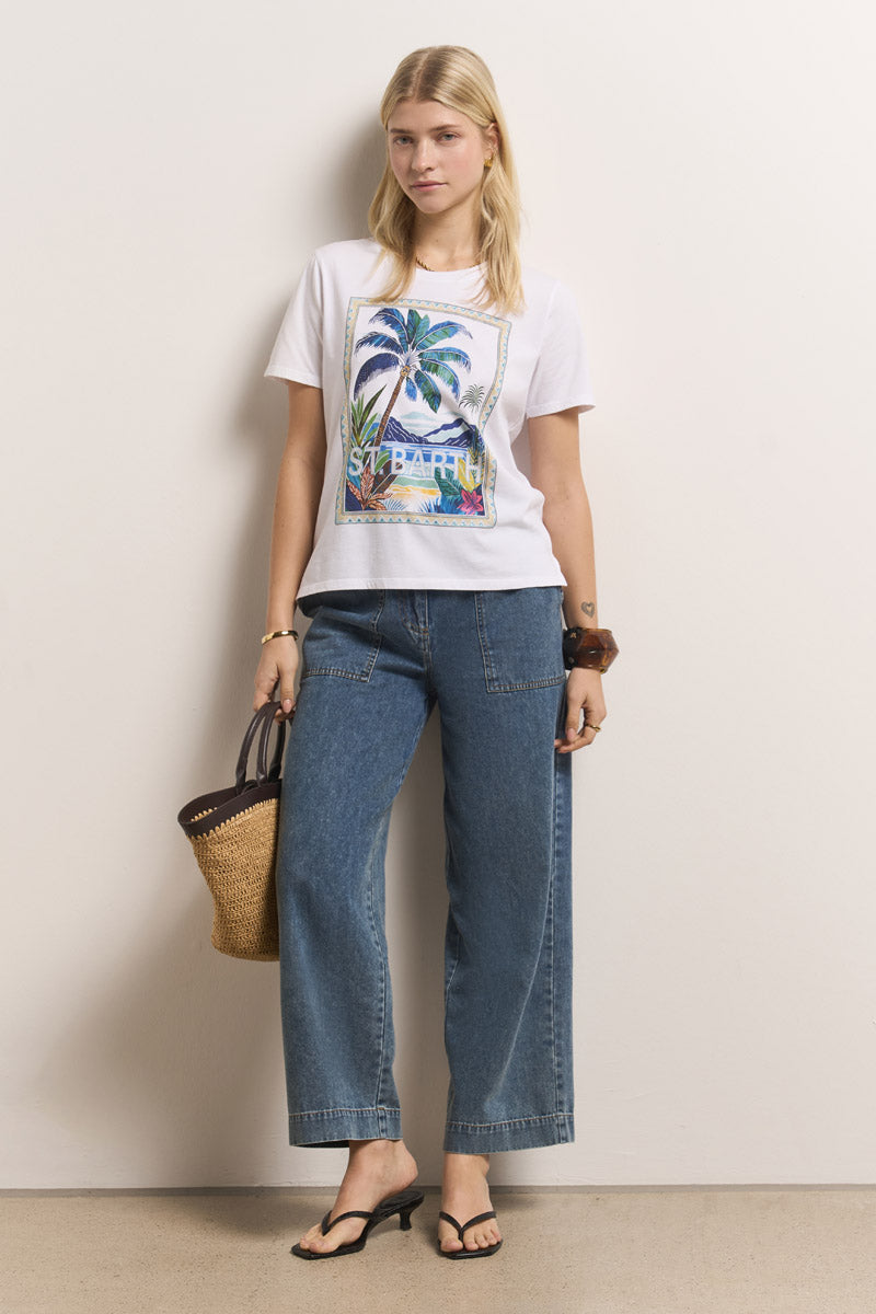 Eine Frau mit glattem blondem Haar trägt ein weißes Princess Goes Hollywood St. Barth T-Shirt aus Baumwolle mit einer Palmengrafik, weite Bluejeans, schwarze Sandalen, hat eine geflochtene Strohtasche in der Hand und trägt ein braunes Haarband am Handgelenk.