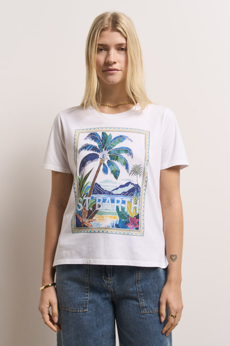 Eine Frau mit glattem blondem Haar trägt ein weißes Baumwolle-T-Shirt von Princess Goes Hollywood mit einer bunten tropischen St. Barth-Grafik, gepaart mit blauen Jeans, Goldschmuck und einer kleinen Herz-Tätowierung auf ihrem linken Unterarm vor einem schlichten Hintergrund.