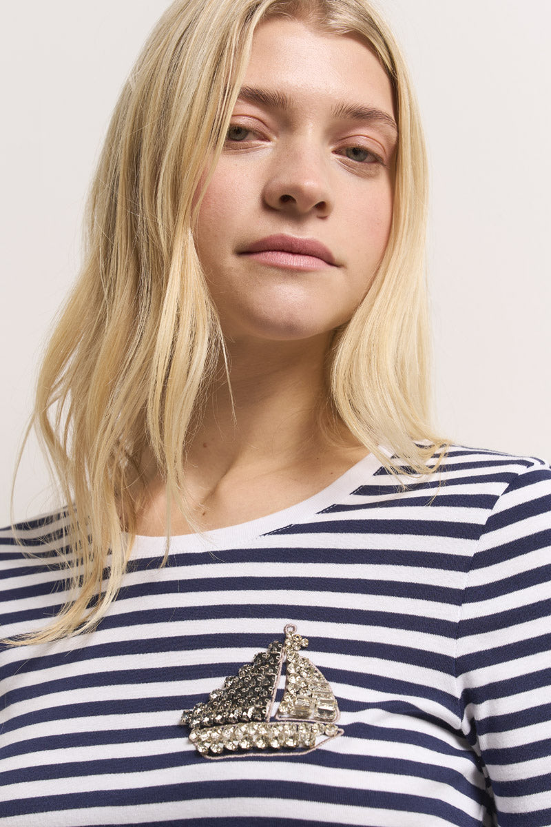 Eine blonde Frau, die ein marineblau-weiß gestreiftes Baumwoll-T-Shirt von Princess Goes Hollywood trägt, blickt ruhig in die Kamera. An ihrer linken Schulter trägt sie eine große, mit Strasssteinen besetzte Schuhbrosche. Der Hintergrund ist schlicht und neutral.