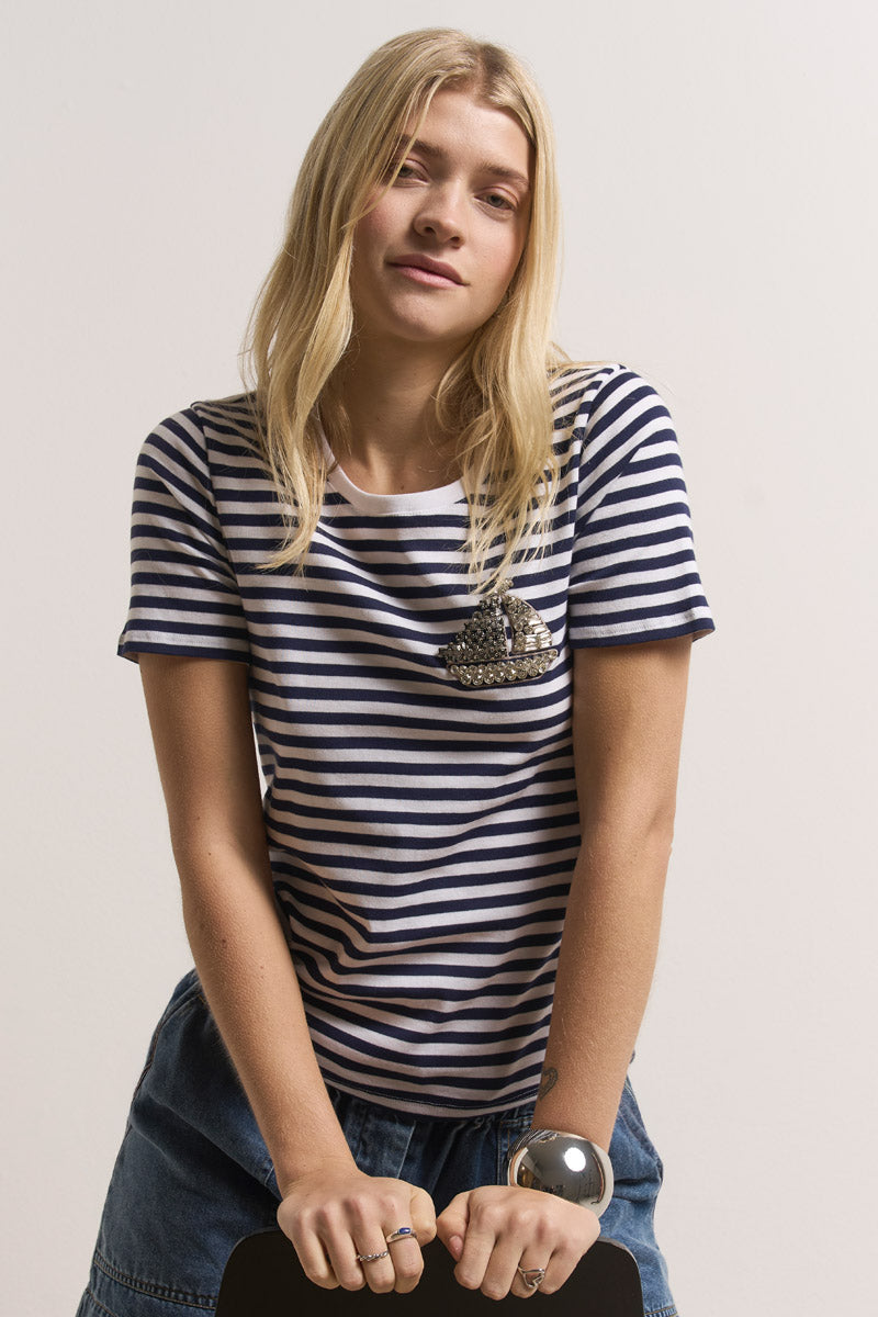 Eine junge Frau mit langen blonden Haaren trägt ein gestreiftes Baumwoll-T-Shirt von Princess Goes Hollywood mit einem mit Juwelen besetzten Stiefelmuster auf der Brust und einen Jeansrock. Sie lehnt sich nach vorne, die Hände auf einen Stuhl gestützt, mit einem neutralen Ausdruck und minimalem Hintergrund.