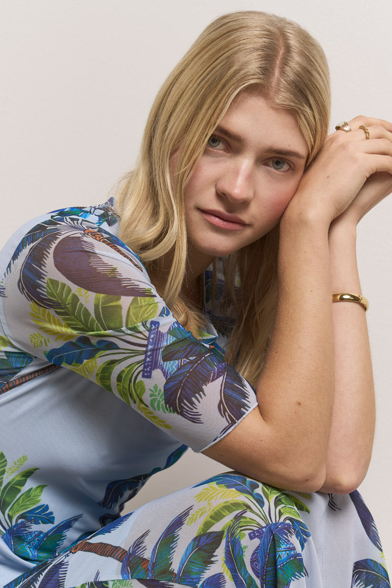 Eine blonde Frau mit heller Haut lehnt sich nach vorne und stützt ihre Arme auf ihre Knie. Sie trägt ein hellblaues Mesh-Kleid mit Palmen-Print von Princess Goes Hollywood, das mit goldenen Ringen und einem Armband verziert ist. Ihr Gesichtsausdruck ist ruhig, der Hintergrund ist gebrochen weiß.