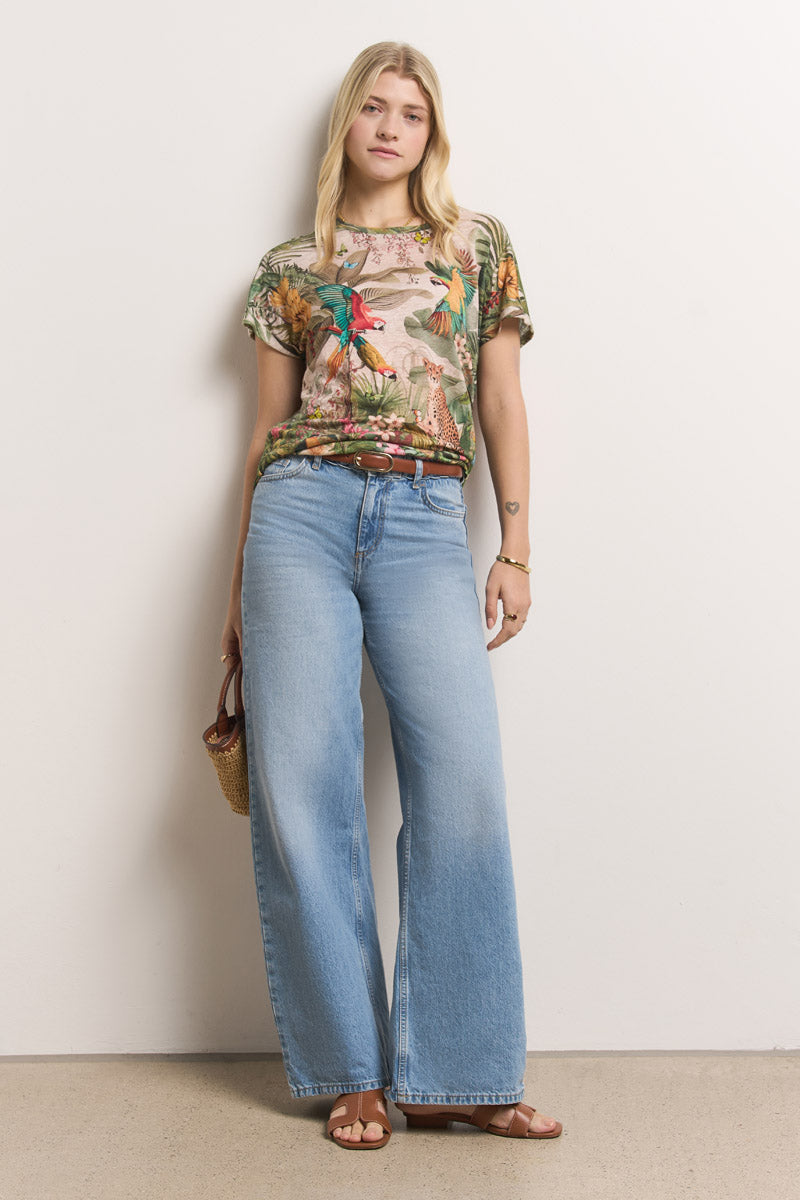 Eine Frau mit langen blonden Haaren steht vor einer weißen Wand. Sie trägt ein tropisch bedrucktes Princess Goes Hollywood Leinen-T-Shirt mit Rundhalsausschnitt, hellblaue Jeans mit hoher Taille und weitem Bein, hellbraune Sandalen und eine Handtasche aus Stoff. Schlichter Schmuck vervollständigt ihren Look.