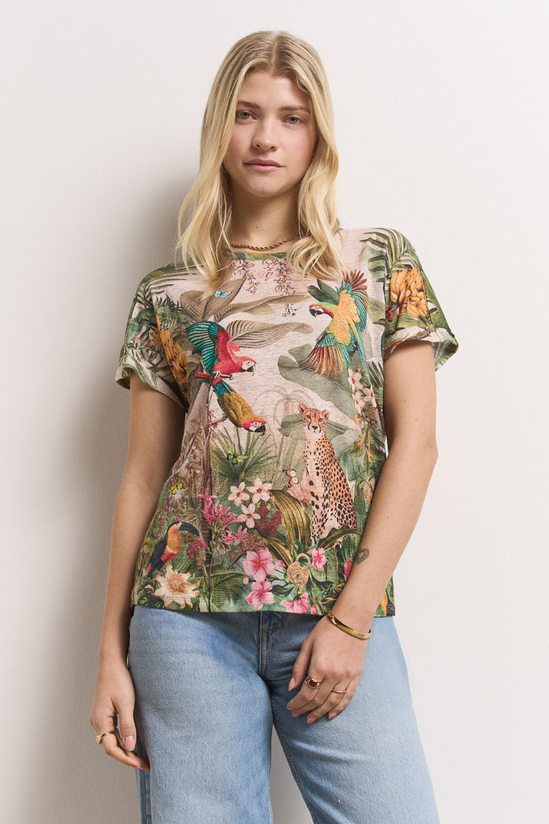 Eine blonde Frau steht vor einer weißen Wand und trägt ein Princess Goes Hollywood T-Shirt aus Leinen mit tropischem Aufdruck und Rundhalsausschnitt, auf dem Papageien, Blumen und ein Gepard abgebildet sind. Sie kombiniert es mit hellblauen Jeans und minimalem Schmuck und sieht entspannt aus.