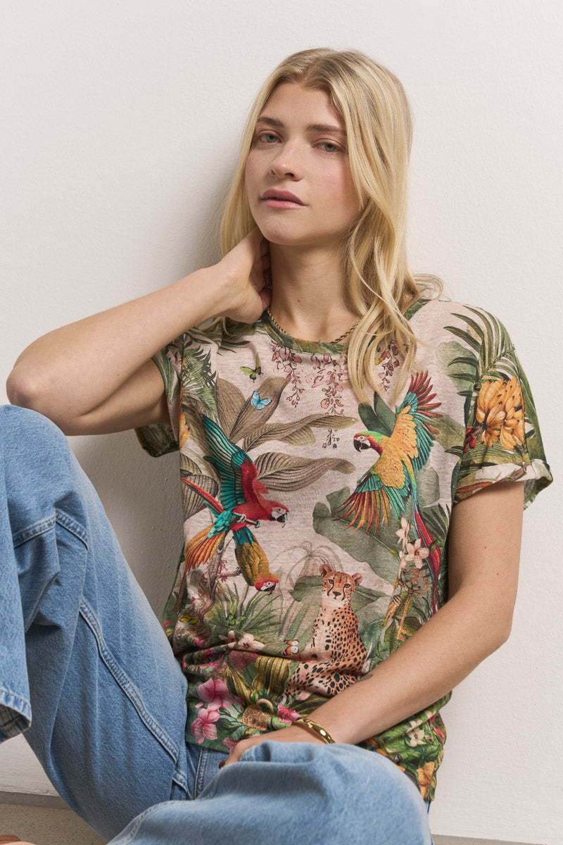 Eine Frau mit langen blonden Haaren sitzt an einer weißen Wand. Sie trägt ein leuchtendes Leinen-T-Shirt mit Dschungeltieren und hellblaue Jeans mit tropischem Aufdruck. Eine Hand berührt ihren Nacken, die andere ruht auf ihrem Knie; sie sieht entspannt und neutral aus.