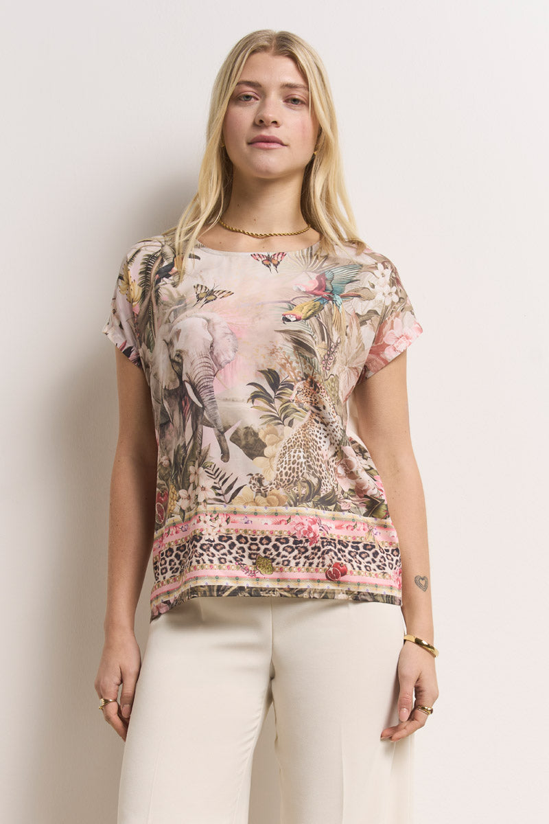 Eine blonde Frau mit einem ruhigen Gesichtsausdruck trägt ein kurzärmeliges, geblümtes T-Shirt mit Rundhalsausschnitt im Jungle-Print von Princess Goes Hollywood, eine cremefarbene Hose, minimalen Schmuck und ein kleines Herz-Tattoo am linken Handgelenk.