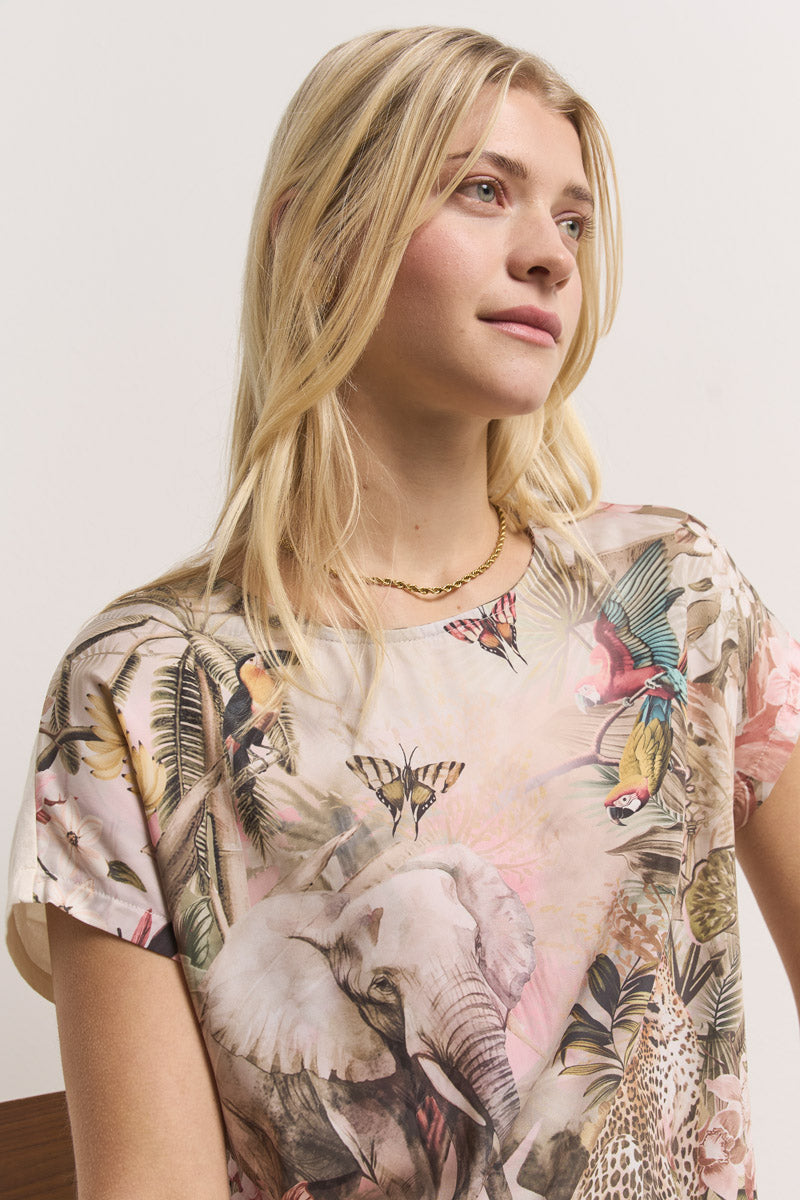 Eine junge Frau trägt ein pastellfarbenes T-Shirt mit Dschungeldruck von Princess Goes Hollywood, auf dem Tiere und Blätter abgebildet sind. Sie blickt ruhig zur Seite und hebt den klassischen Rundhalsausschnitt ihres Oberteils hervor.