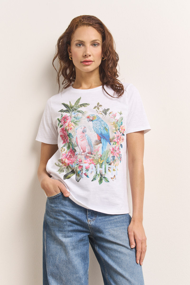 Eine selbstbewusste Frau mit gewelltem kastanienbraunem Haar modelliert ein Princess Goes Hollywood T-Shirt aus Baumwolle mit lebhaften Blumendrucken, zwei Papageienmotiven und dem Text "TAHITI". Sie posiert vor einer hellen Wand, die Hand in der Tasche ihrer Blue Jeans.