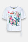 Princess Goes Hollywood weißes kurzärmeliges T-Shirt aus Baumwolle mit einem bunten Tukan zwischen rosa Tropenblumen und grünen Blättern, "Paradise" in roter Schrift über dem Vogel und einem Kordelzug am unteren Saum.